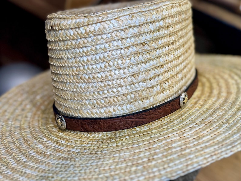 May include: A straw hat with a wide brim and a cylindrical crown. The hat features a dark brown leather band with a floral pattern and silver-colored decorative studs. The hat is designed for sun protection and outdoor wear.