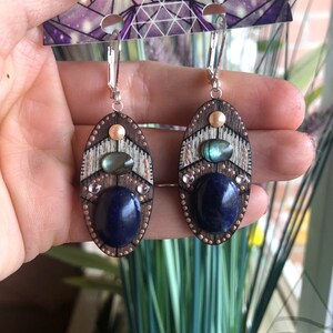 May include: A pair of wooden earrings with a geometric design. The earrings feature a blue lapis lazuli stone, a white mother of pearl shell, and a copper accent. The earrings are hanging from silver hooks.