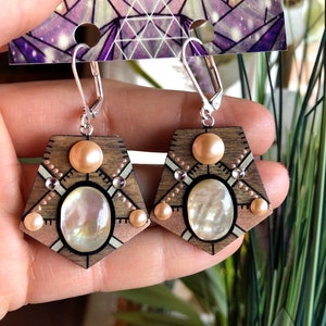 Op de afbeelding: Een paar houten oorbellen met een geometrisch ontwerp. De oorbellen zijn gemaakt van hout en hebben een zilveren haak. De oorbellen zijn versierd met een witte schelp, roze parels en koperen accenten. De oorbellen zijn gelabeld "Temple Jewels".