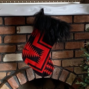 Puede incluir: Una media roja y negra con un patrón geométrico cuelga de una repisa blanca sobre una chimenea de ladrillo. La media tiene un puño de piel sintética negra.