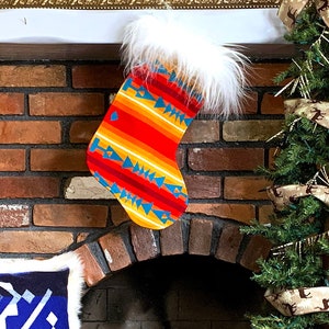 Puede incluir: Una chimenea de ladrillo con una media de punto de imitación de piel blanca con un patrón geométrico colorido. La media está colgada en la repisa de la chimenea sobre una chimenea con una rejilla de metal negro y troncos. Hay dos pequeños árboles de Navidad decorados con cinta marrón y azul a cada lado de la chimenea.