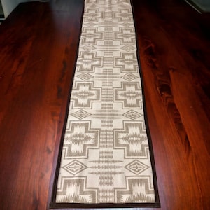 Puede incluir: Camino de mesa rectangular con un patrón geométrico en beige y marrón. El patrón presenta formas de cruz y diseños de diamantes. El camino de mesa está sobre una mesa de madera marrón oscuro y tiene un borde marrón oscuro.