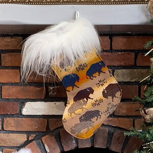 Peut inclure: Une chaussette de Noël avec un motif beige et jaune représentant des silhouettes de bisons. La chaussette a un revers en fausse fourrure blanche et est suspendue à une cheminée en briques. Le motif comprend des accents marron, bleu et orange.