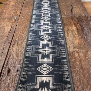 Op de afbeelding: Zwarte en witte geometrische tafelrunner met een zwarte leren bies. De runner heeft een zuidwestelijk ontwerp.