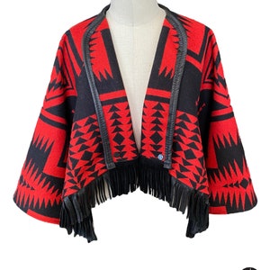 May include: A red and black patterned jacket with a fringed hem. The jacket features a geometric design and black leather trim along the collar and edges. The jacket is displayed on a mannequin.