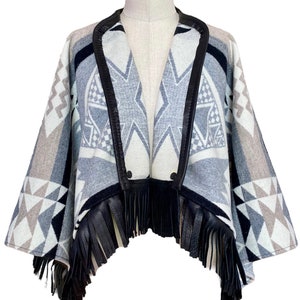 May include: A gray and black patterned poncho with a black leather trim and fringe. The poncho has a geometric design and is made of a soft, warm fabric.
