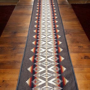 Op de afbeelding: Een lange, rechthoekige tafelrunner met een geometrisch patroon in tinten bruin, wit, rood, oranje en blauw. De runner heeft een zwarte rand.