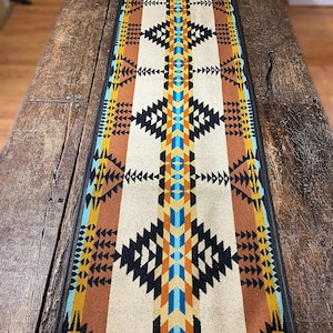Puede incluir: Un camino de mesa blanco con un patrón geométrico en tonos de marrón, negro, azul y amarillo. El camino de mesa tiene un borde negro y está hecho de un tejido suave y tejido.