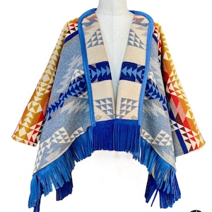 May include: A blue and beige patterned poncho with a blue suede fringe. The poncho has a V-neck and is made of a soft, woven fabric.