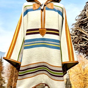 Könnte beinhalten: Ein weißer Kapuzenponcho mit braunen Lederbesätzen und einem gestreiften Muster in den Farben Gelb, Orange, Blau, Grün und Braun.