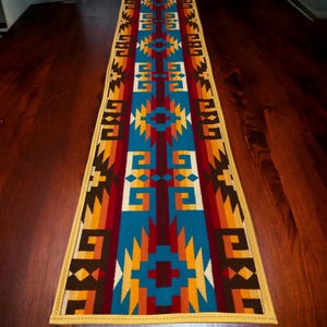 Puede incluir: Un camino de mesa rectangular con un diseño del suroeste. El camino de mesa presenta un patrón repetitivo de formas geométricas en tonos de azul, rojo, amarillo y marrón. Los bordes están ribeteados con un borde amarillo. El camino de mesa está colocado sobre una mesa de madera oscura.