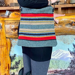 May include: A woman wearing a black sweater and black leggings is carrying a gray and red striped crossbody bag. The bag has a black strap and is made of a soft, woven material. The bag is worn over the shoulder and is hanging down the side of the woman's body.