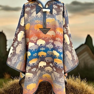Peut inclure: Un poncho à capuche avec une bordure en cuir marron et un motif de buffle dans des tons de marron, bleu et orange. Le poncho est présenté sur une botte de foin.