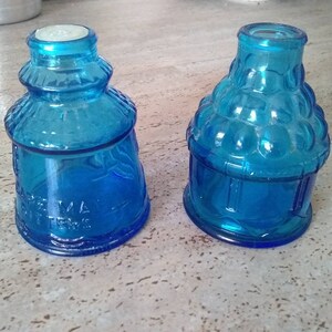 May include: Two blue glass bottles with different designs. The bottle on the left is taller and has a wider base. The bottle on the right is shorter and has a more rounded shape. Both bottles have a blue tint and are made of glass.