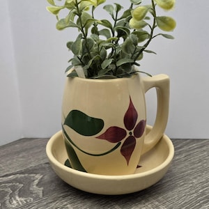 May include: A cream-colored ceramic mug with a matching saucer, featuring a painted dark red four-petaled flower and green leaves. The mug contains a small artificial green plant with rounded leaves, creating a decorative botanical display.