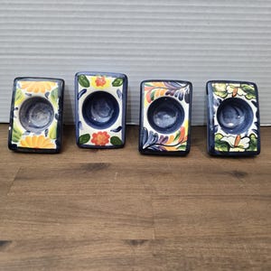 Puede incluir: Cuatro candelabros rectangulares de cerámica con diseños florales. Cada soporte tiene un interior azul oscuro y está decorado con flores y hojas coloridas. Los soportes están dispuestos sobre una superficie de madera.