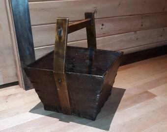 Puede incluir: Un cubo de madera marrón oscuro con un asa de latón. El cubo está sentado en un suelo de madera clara.