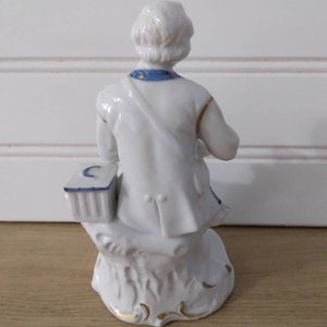 Puede incluir: Figurilla de porcelana blanca de una persona sentada en un toc&oacute;n de &aacute;rbol. La persona lleva una camisa con estampado azul y blanco y un sombrero blanco. La figurita tiene detalles dorados.