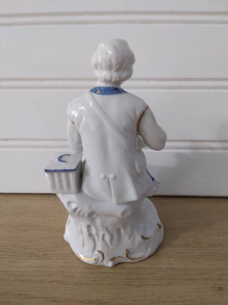 Puede incluir: Figurilla de porcelana blanca de una persona sentada en un toc&oacute;n de &aacute;rbol. La persona lleva una camisa con estampado azul y blanco y un sombrero blanco. La figurita tiene detalles dorados.