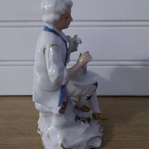 Puede incluir: Una figurita de porcelana de una persona con un atuendo blanco y azul, sentada en una roca con detalles dorados. La figurita est&aacute; en posici&oacute;n sentada, sosteniendo un objeto peque&ntilde;o en su mano.