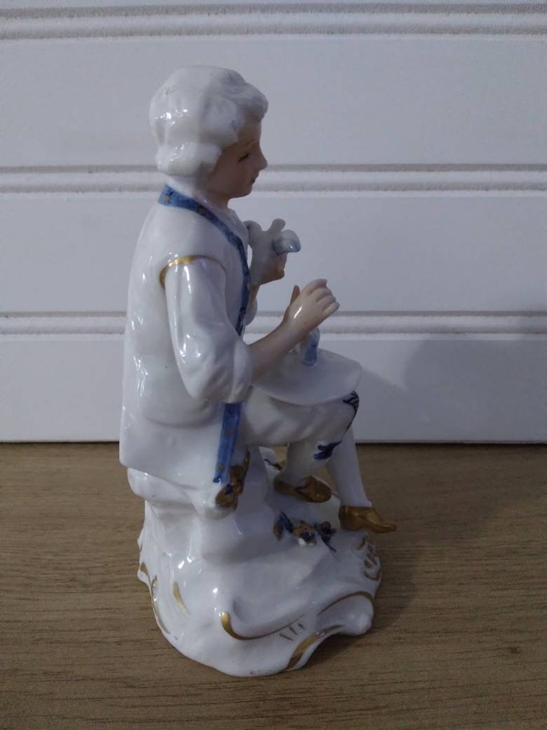 Puede incluir: Una figurita de porcelana de una persona con un atuendo blanco y azul, sentada en una roca con detalles dorados. La figurita est&aacute; en posici&oacute;n sentada, sosteniendo un objeto peque&ntilde;o en su mano.