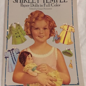May include: A vintage paper doll book featuring Shirley Temple. The cover shows a color illustration of Shirley Temple holding a doll. The book title is "Classic Shirley Temple Paper Dolls in Full Color" with the text "Edited by Grace Piamontesi".
