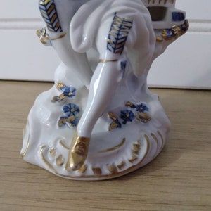 Puede incluir: Una figurita de porcelana de una persona sentada en una roca con acentos azules y dorados. La figurita es blanca con detalles azules y dorados.