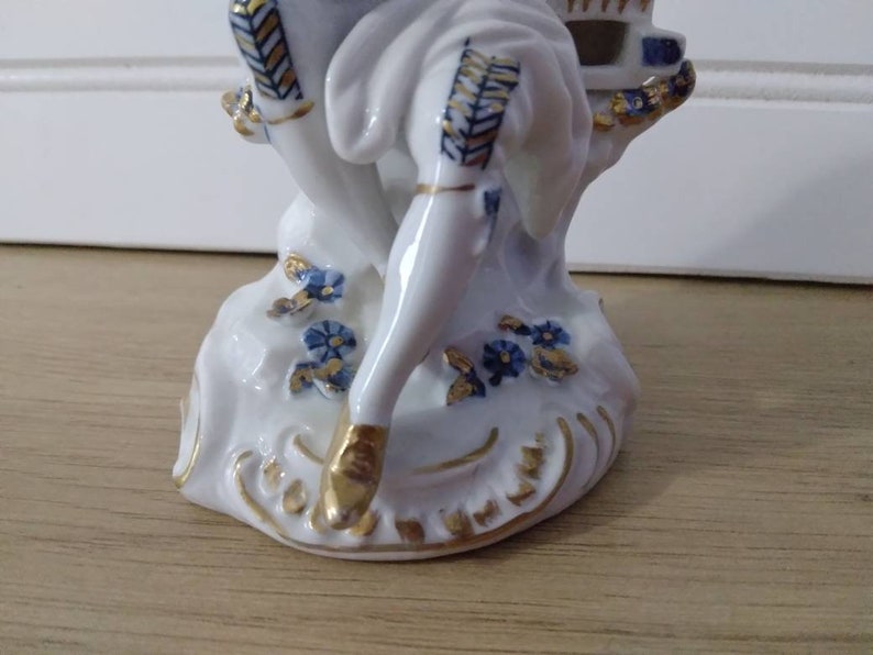 Puede incluir: Una figurita de porcelana de una persona sentada en una roca con acentos azules y dorados. La figurita es blanca con detalles azules y dorados.