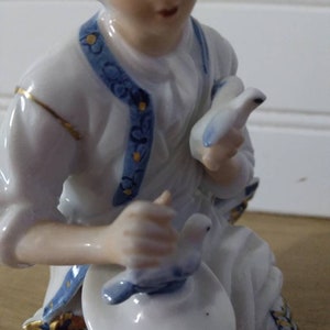 Puede incluir: Una figurita de porcelana de una persona sosteniendo dos palomas blancas con detalles azules. La figurita lleva un atuendo estampado en blanco y azul con detalles dorados.