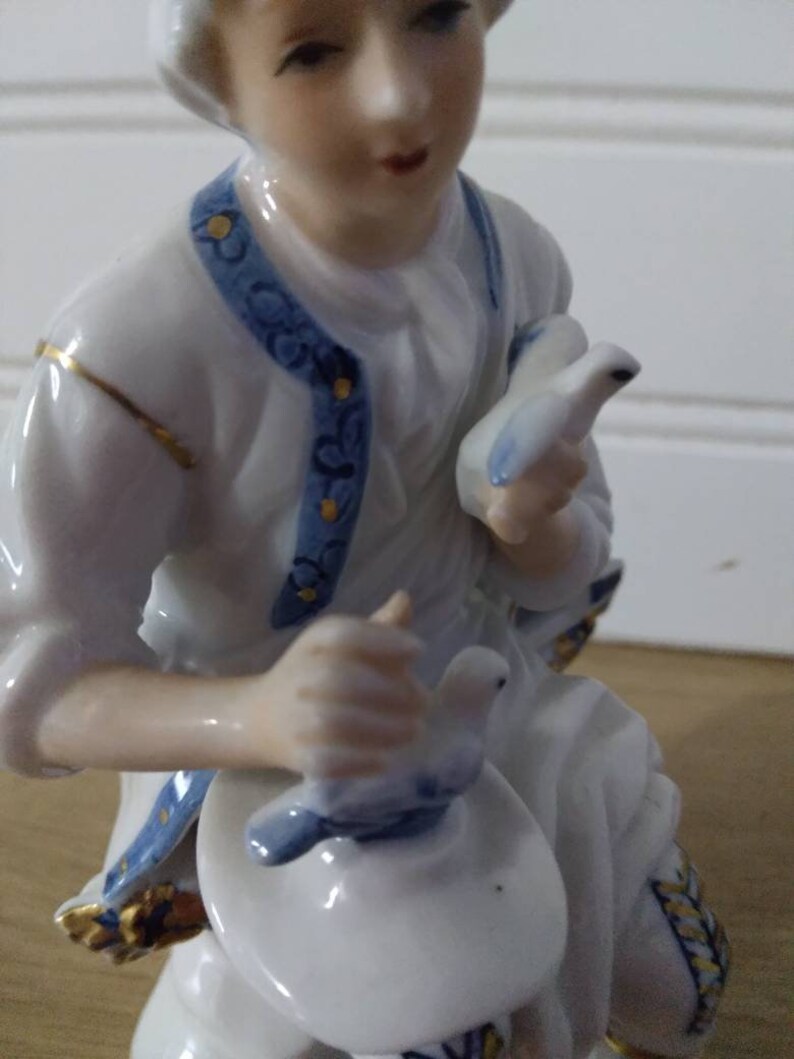Puede incluir: Una figurita de porcelana de una persona sosteniendo dos palomas blancas con detalles azules. La figurita lleva un atuendo estampado en blanco y azul con detalles dorados.