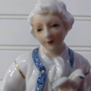 Puede incluir: Una figurita de porcelana de una persona que lleva una chaqueta con estampado azul y blanco con ribetes dorados. La figurita tiene una peluca blanca y un cuello blanco.