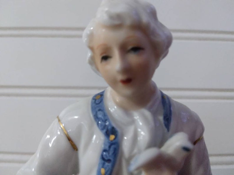Puede incluir: Una figurita de porcelana de una persona que lleva una chaqueta con estampado azul y blanco con ribetes dorados. La figurita tiene una peluca blanca y un cuello blanco.