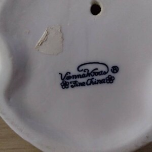 Puede incluir: Figurita de porcelana blanca con una inscripci&oacute;n azul y negra que dice "Vienna Woods Fine China" con un peque&ntilde;o dise&ntilde;o de flores a cada lado del texto.