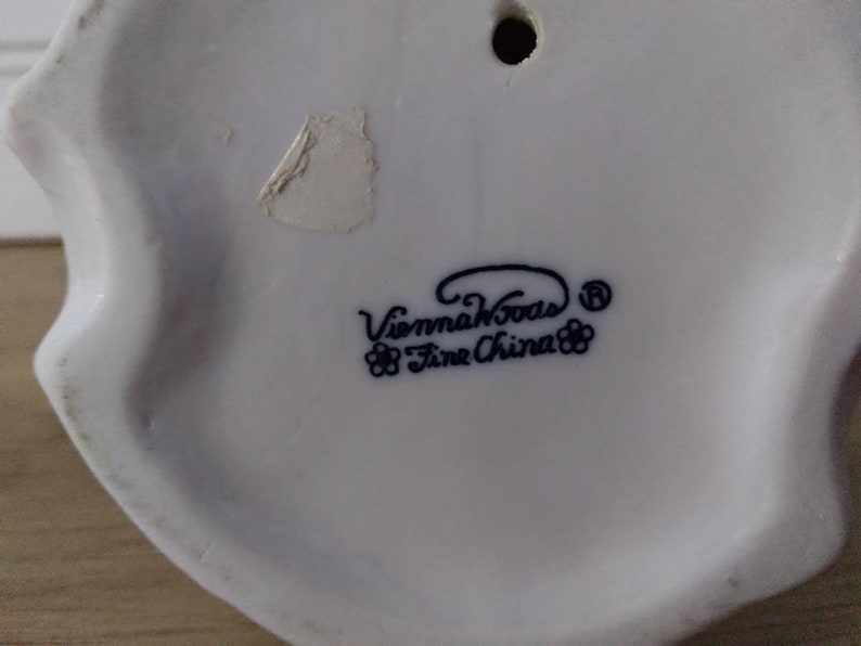 Puede incluir: Figurita de porcelana blanca con una inscripci&oacute;n azul y negra que dice "Vienna Woods Fine China" con un peque&ntilde;o dise&ntilde;o de flores a cada lado del texto.