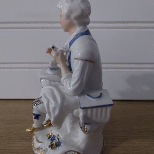 Puede incluir: Una figurita de porcelana de una persona sosteniendo un p&aacute;jaro. La figurita est&aacute; pintada en blanco, azul y dorado. La persona lleva una camisa blanca con detalles azules y un sombrero azul y blanco. El p&aacute;jaro es blanco con detalles azules. La figurita est&aacute; sentada sobre una base con dise&ntilde;os florales azules y dorados.