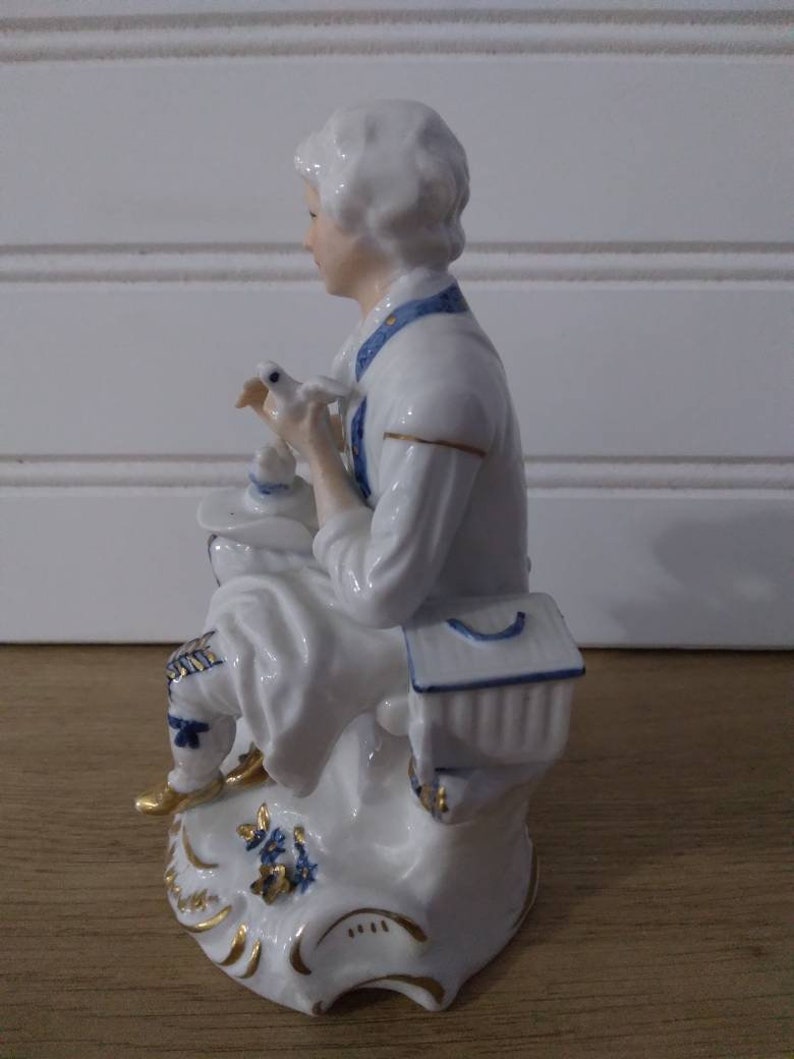 Puede incluir: Una figurita de porcelana de una persona sosteniendo un p&aacute;jaro. La figurita est&aacute; pintada en blanco, azul y dorado. La persona lleva una camisa blanca con detalles azules y un sombrero azul y blanco. El p&aacute;jaro es blanco con detalles azules. La figurita est&aacute; sentada sobre una base con dise&ntilde;os florales azules y dorados.