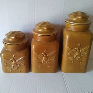 Könnte beinhalten: Set aus drei braunen Keramikbehältern mit Adler- und Sternmotiven. Die Behälter haben Deckel und sind perfekt zum Aufbewahren von Küchenutensilien.