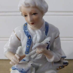 Puede incluir: Una figurita de porcelana de un joven que sostiene una paloma en su mano. La figurita lleva una camisa blanca con ribetes azules y dorados. La paloma est&aacute; pintada en tonos de azul y blanco.