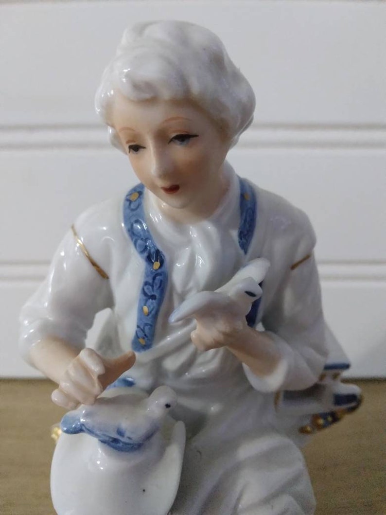 Puede incluir: Una figurita de porcelana de un joven que sostiene una paloma en su mano. La figurita lleva una camisa blanca con ribetes azules y dorados. La paloma est&aacute; pintada en tonos de azul y blanco.