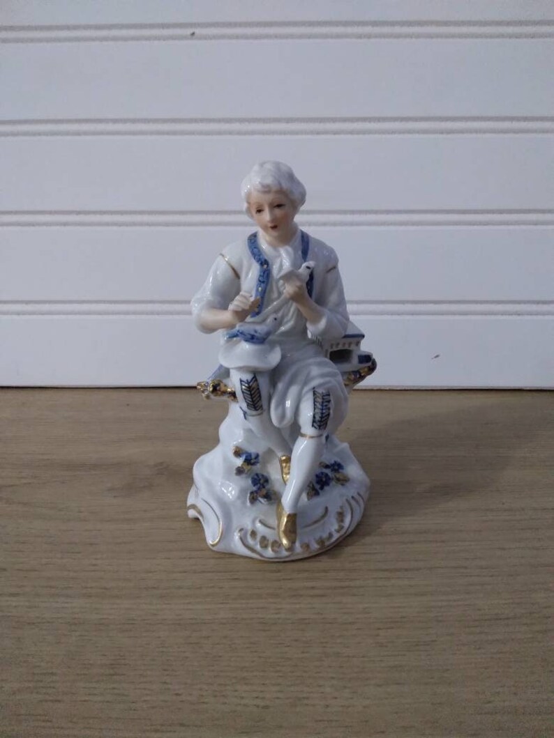 Puede incluir: Una figurita de porcelana de una persona sentada en una base con detalles azules y dorados. La persona lleva una camisa blanca con un chaleco estampado en azul y dorado. La persona sostiene un peque&ntilde;o p&aacute;jaro blanco en su mano.