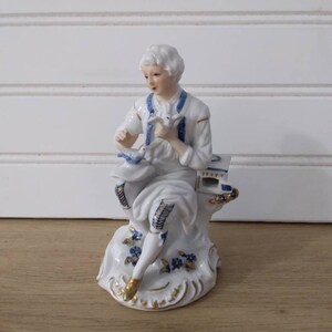 Puede incluir: Una figurita de porcelana de una persona sentada en una roca, sosteniendo un p&aacute;jaro en su mano. La figurita est&aacute; pintada en blanco, azul y dorado.