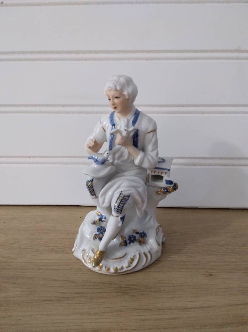 Puede incluir: Una figurita de porcelana de una persona sentada en una roca, sosteniendo un p&aacute;jaro en su mano. La figurita est&aacute; pintada en blanco, azul y dorado.