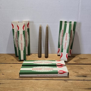 Puede incluir: Un surtido de cajas verdes y blancas de velas perfumadas Bayberry, con dos velas cónicas marrones en el centro. Las cajas tienen un diseño rojo y blanco y el texto "Bayberry Candles". También es visible el texto "The Candle that says Season's Greetings".