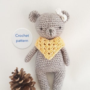 Op de afbeelding: Een gehaakte grijze teddybeer met een geel gehaakt slabbetje en een witte strik. De beer staat op een wit oppervlak met een dennenappel links. De tekst "Crochet pattern" staat op een witte cirkel links van de beer. De tekst "Frisian Knitting" staat onder de beer.