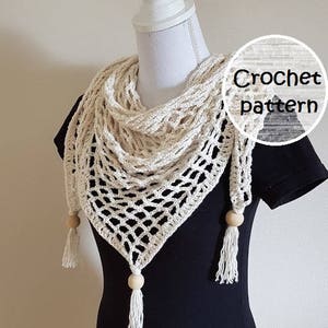 Op de afbeelding: Een gehaakte sjaal met een delicaat opengewerkt patroon in een crèmekleur. De sjaal heeft houten kralen en kwastjes. De tekst "Crochet pattern" en "Frisian Knitting" is zichtbaar in de afbeelding.