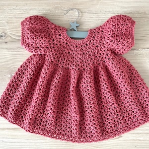 Op de afbeelding: Een handgemaakte, stoffig roze gehaakte jurk voor een baby of peuter. De jurk heeft een ronde halslijn, korte mouwen en een uitlopende rok. Het wordt getoond op een lichtblauwe hanger met een stervormig detail.