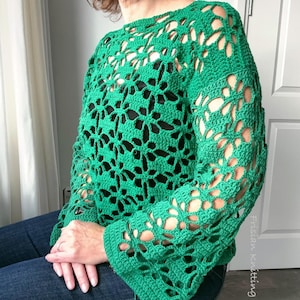 Op de afbeelding: Een groene gehaakte top met een bloemenpatroon. De top met lange mouwen heeft een los, open geweven ontwerp. De mouwen zijn uitlopend bij de pols. De top wordt over een zwart kledingstuk gedragen.