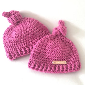 Op de afbeelding: Twee roze gebreide mutsen met een knoop bovenop. Een muts heeft een klein houten label met de naam "anna" erop.