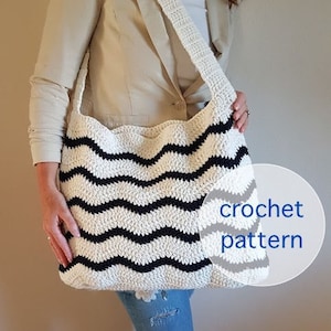 Op de afbeelding: Een witte en zwarte gestreepte gehaakte tote bag met een lange band. De tas wordt over de schouder gedragen door iemand die een beige blazer en een blauwe jeans draagt. De tekst "crochet pattern" staat op de tas.