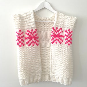 May include: Cream-colored, sleeveless knitted vest with a chunky knit texture. The vest features bright pink geometric patterns on the shoulders. The garment is displayed on a white hanger against a white background.